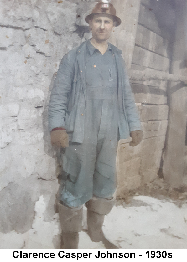 Colorized photo of Clarence Casper Johnson, with heavy 5 o'clock shadow, wearing a mining helmet with light, heavy work overalls and jacket, and heavy gloves and knee boots, standing on white sand against a rock wall.